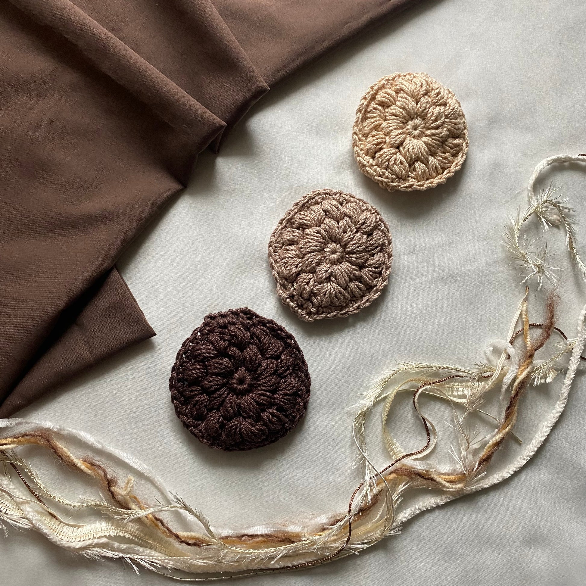 Three crocheted coasters in brown and beige on a marble surface with fabric and ribbons.