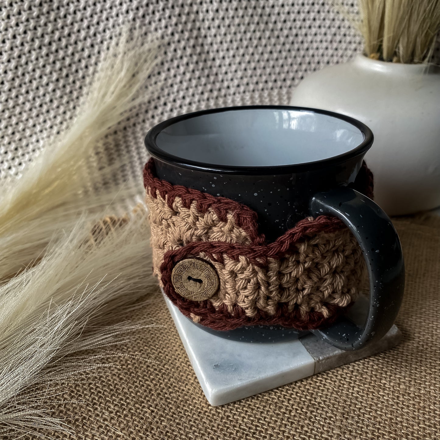 Black mug with a brown and beige crocheted cozy on a marble coaster, with a textured background.