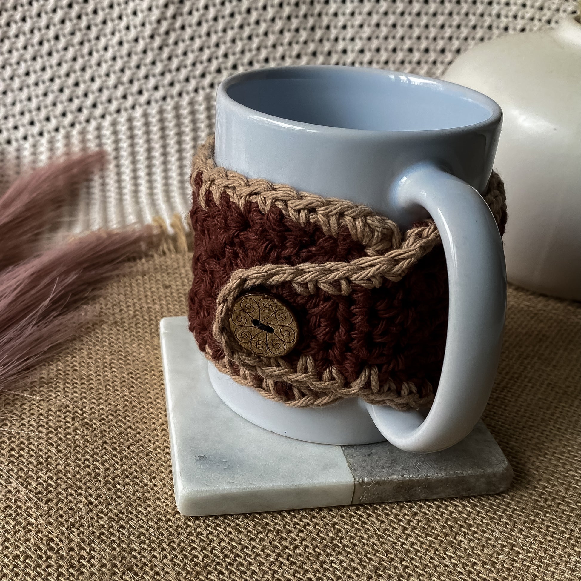 White mug with a brown crochet cozy on a marble coaster, textured background