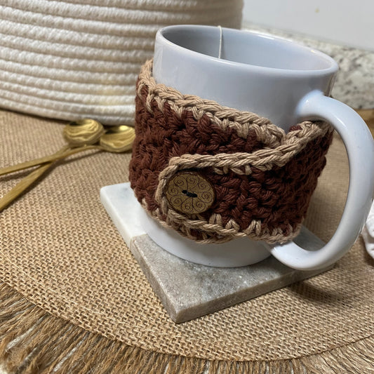 White mug with a brown crochet cozy on a marble coaster, placed on a textured surface.