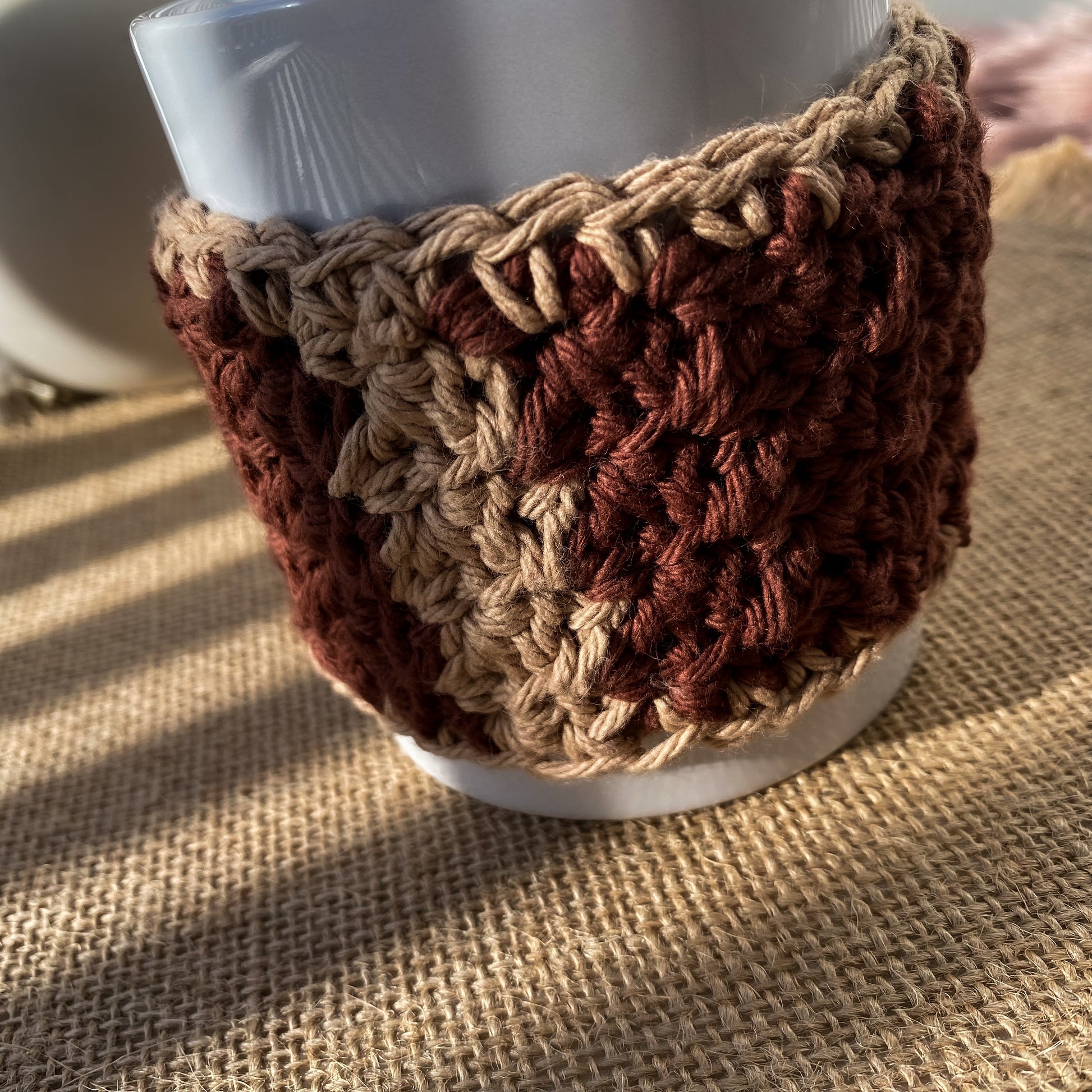 Crocheted brown and beige cup cozy on a white mug with another similar mug in the background.